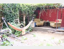 Ein kleiner Gartenbereich mit einer Hängematte im Vordergrund, umgeben von Pflanzen und Efeu bewachsenen Strukturen. Im Hintergrund befinden sich zwei Sessel und ein kleiner Tisch unter einer überdachten Pergola mit roten Backsteinwänden.