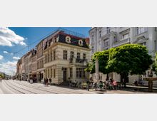 Straßenszene mit historischen Gebäuden und Außencafé: Ein zweistöckiges Eckgebäude mit dekorativen Fassadenelementen und schwarzen Mansardendächern steht neben weiteren Altbauten. Vor dem Gebäude befindet sich ein Café mit Sitzplätzen unter schattenspendenden Bäumen. Im Vordergrund verlaufen Straßenbahnschienen entlang der gepflasterten Straße.