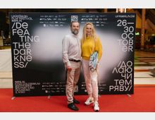Ein Mann und eine Frau stehen auf einem roten Teppich vor einer schwarzen Plakatwand mit der Aufschrift „Defeating the Darkness“ und weiteren Informationen zum Ukrainian Film Festival Berlin, das vom 26. bis 30. Oktober stattfindet. Im Hintergrund sind ein heller Innenbereich und Treppen zu sehen.