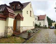 Zwei helle Wohnhäuser mit roten Ziegeldächern und verputzten Fassaden, verbunden durch einen gepflasterten Weg. Das vordere Haus zeigt dekorative Mauerdetails aus Naturstein und roten Ziegeln an der Eingangstür, daneben ein Blumentopf mit weißen Blumen.