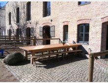 Backsteinfassade eines Gebäudes mit mehreren großen Fenstern und gewölbten Fensterrahmen, davor ein gepflasterter Hof mit einem langen Holztisch und Bänken. Holzgeländer grenzen den Bereich ab, und im Hintergrund ist ein Strauch zu sehen.
