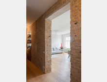 Wohnraum in einem modernen Apartment mit freigelegten Ziegelwänden, Holzboden und einer offenen Türöffnung. Im Hintergrund steht ein graues Sofa vor einer weißen Wand neben einem großen Fenster mit Tageslicht.