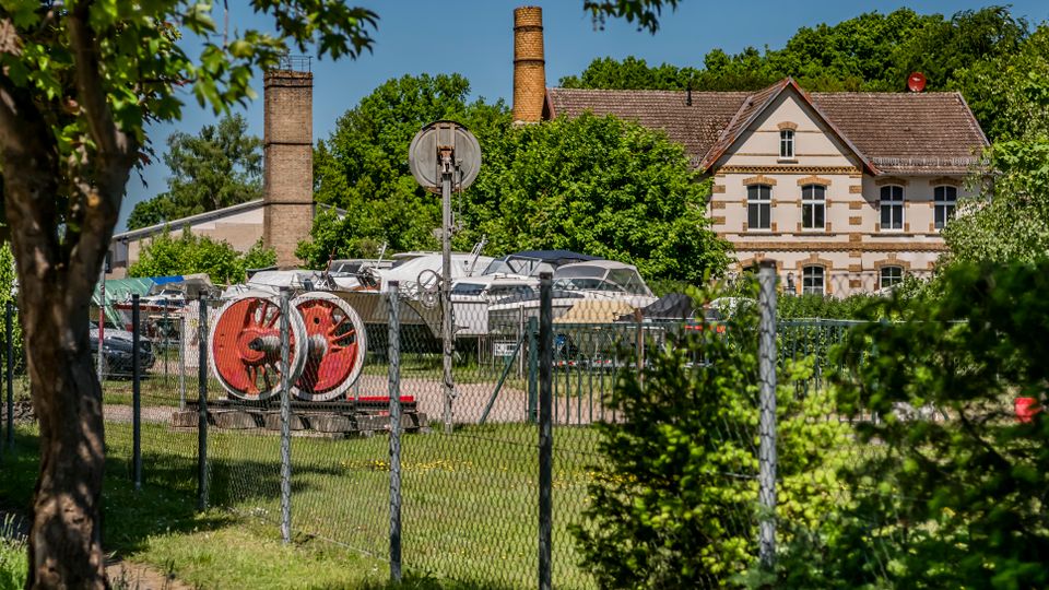 Eingezäuntes Industrie- und Lagergelände mit Booten und Geräten im Freien, im Hintergrund zwei hohe Backsteinschornsteine. Rechts steht ein zweigeschossiges, historisch wirkendes Gebäude mit hellem Putz, Ziermauerwerk und Satteldach, umgeben von dichtem Grün.