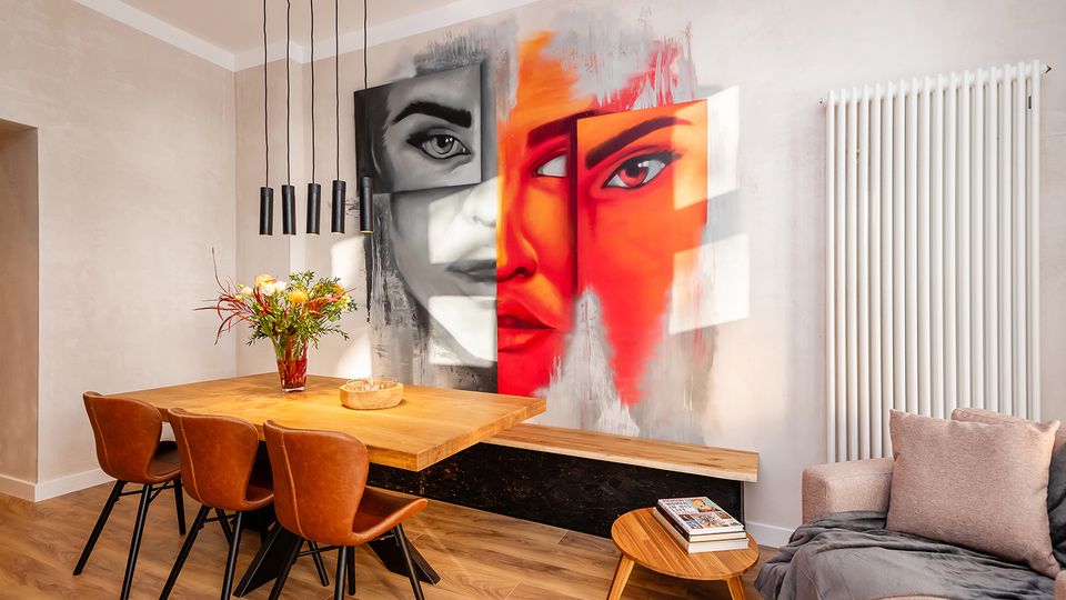 Moderner Ess- und Wohnbereich mit Holztisch, braunen Stühlen und hängenden schwarzen Pendelleuchten, kombiniert mit einem Sofa und kleinem Couchtisch. An der Wand dominiert ein großes, farbiges Wandgemälde mit abstrakten Gesichtern; daneben steht ein hoher, weißer Heizkörper.