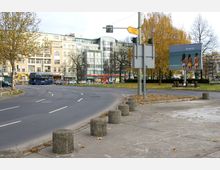Straßenkreuzung in einer städtischen Umgebung mit mehrspuriger Fahrbahn, umgeben von Bäumen mit Herbstlaub, Wohn- und Geschäftsgebäuden im Hintergrund. Rechts befindet sich eine Werbetafel, daneben ein Wegweiser und Poller am Rand des Gehwegs.
