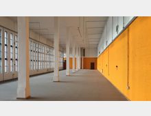 Leerer, rechteckiger Innenraum mit hohen Decken, weißen Pfeilern und einer langen Fensterreihe auf der linken Seite. Die rechte Wand ist in einem kräftigen Gelb gestrichen, während sichtbare Leitungen und Lampen die industrielle Atmosphäre unterstreichen.