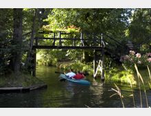 Zwei Personen paddeln in einem Kanu auf einem ruhigen Fluss unter einer schmalen Holzbrücke hindurch. Die Umgebung ist von dichtem Grün geprägt, mit blühenden Pflanzen und Bäumen im Hintergrund.