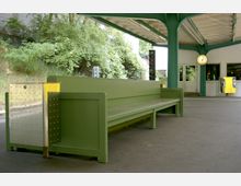 Grüner Bahnsteigbereich mit Sitzbank aus Holz und Metallabfallbehälter; im Hintergrund eine Bahnhofsuhr, Türen und ein gelber Fahrkartenautomat, umgeben von Bäumen und einer weißen Gebäudeoberfläche.