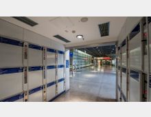 Ein moderner Bahnhofsbereich mit Schließfächern an den Seitenwänden, einem glatten Boden und einer offenen Durchgangshalle. Im Hintergrund befinden sich Rolltreppen, Glaswände und Beschilderungen, die zu weiteren Bereichen führen.