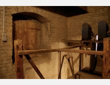 Ein rustikaler Raum mit groben Steinmauern und Holzstrukturen, darunter ein Holztor in einem gewölbten Mauerdurchgang. Im Hintergrund sind zwei blaue mechanische Geräte sichtbar, die möglicherweise Teil einer alten technischen Anlage sind.