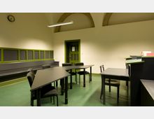 Ein schlichter Seminarraum mit grünem Boden und weißen Wänden. Mehrere schwarze Tische und Stühle stehen im Raum, an den Wänden befinden sich grüne Tafelflächen, und eine grüne Tür führt aus dem Raum.