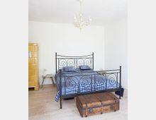 Ein schlichter Schlafzimmerraum mit einem schwarzen Metallbett mit geschwungenen Verzierungen, blauer karierten Bettwäsche, einem gelben Kachelofen links und einer antiken Holzkiste am Fußende des Bettes. Die Wände sind weiß und die Decke trägt einen dekorativen Kronleuchter.