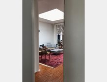 Blick in ein modernes Wohnzimmer mit heller Einrichtung, einem grauen Sofa auf einem großen roten Orientteppich, Holzboden sowie einer Deckenöffnung, die Tageslicht hereinlässt. Ein Kristalllüster hängt mittig im Raum und dekorative Möbel wie Stühle und ein Couchtisch ergänzen die Einrichtung.