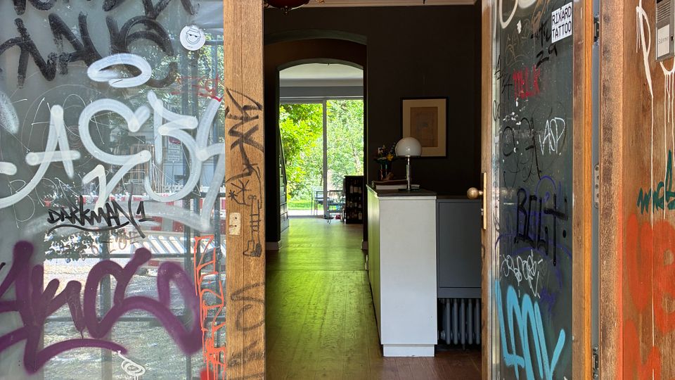 Offene Eingangstür eines Gebäudes mit stark beschmierten Glasflächen und Holzrahmen, die in einen schmalen Flur mit Holzboden führt. Im Inneren sind eine Deckenleuchte und ein weißer Tresen zu sehen; am Ende des Flurs öffnet sich der Blick zu einem hellen Bereich mit großen Fenstern und Grün draußen.
