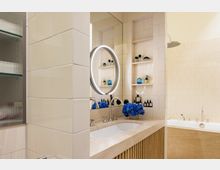 Modernes Badezimmer mit beigefarbenen, gefliesten Wänden. Im Vordergrund ein breites Waschbecken mit einer Steinoberfläche, darüber ein runder beleuchteter Spiegel; daneben blaue Blumen und Pflegeprodukte. Im Hintergrund eine eingelassene Badewanne und Regale mit weiteren Produkten.