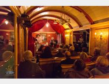 Innenansicht eines kleinen Theaters mit gebogenem Holzgewölbe, roten Samtvorhängen und warmem Licht. Eine Frau in Kostüm steht auf der Bühne, während das Publikum auf Holzbänken sitzt und zuschaut.