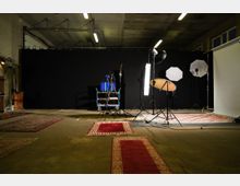 Foto eines Fotostudios mit einem schwarzen Hintergrund. Im Vordergrund sind mehrere rote Teppiche auf einem Betonboden ausgelegt, während im Hintergrund verschiedene Studiobeleuchtungen, Reflektoren und ein Kamerastativ aufgestellt sind.