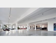 Großer, heller Raum in einem modernen Gebäude mit weiß gestrichenen Wänden, freiliegenden Pfeilern und einer Betondecke. Links befinden sich Sitzbereiche mit Pflanzen und roten Sofas, rechts eine offene, mit dunklen Fliesen verkleidete Küchenzeile; große Fenster sorgen für viel Tageslicht.