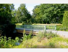 Eine grüne Holzbrücke über einen schmalen Wasserlauf, umgeben von üppiger Vegetation und großen Bäumen in einer Parklandschaft. Der Weg neben der Brücke ist mit Kies bedeckt, und die Umgebung wirkt naturnah und ruhig.