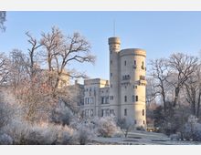Historische Burg mit runden Türmen und Zinnen aus sandfarbenem Stein, umgeben von winterlicher Vegetation mit frostbedeckten Bäumen und Sträuchern unter klarem, blauem Himmel.