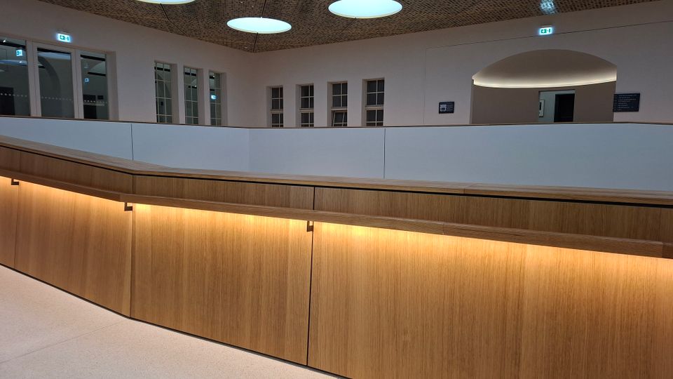 Innenansicht eines modernen Foyers oder Flurs mit geschwungener Brüstung aus Holz und indirekter Beleuchtung. Helle Wände mit mehreren Fenstern, ein großer Rundbogen-Durchgang sowie runde Deckenleuchten unter einer perforierten Decke prägen den Raum.