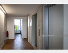 Ein Innenflur mit Holzfußboden, einer geschlossenen Aufzugstür auf der rechten Seite und einer Tür am Ende des Gangs, durch die Tageslicht einfällt. An der Wand hängen reflektierende Sicherheitswesten, und ein Feuerlöscher ist links angebracht.