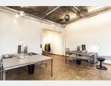 Moderner Büroraum mit weiß gestrichenen Wänden, freiliegender Betondecke und Industrieleuchten. Der Raum ist mit großen, grau gefassten Tischen, Bürostühlen, Computern und Schreibtischlampen ausgestattet; im Hintergrund befindet sich ein Kleiderständer mit Jacken.