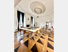 Heller Konferenzraum mit U-förmig angeordneten Tischen und Holzstühlen auf einem geometrisch gemusterten Parkettboden. Hohe Fenster mit dunklen Vorhängen und eine auffällige, spiralförmige Stuckdecke mit Hängelampe prägen den Raum; seitlich befindet sich eine Tür zu einem angrenzenden Bereich.