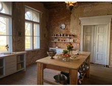 Rustikale Küche mit unverputzten Backsteinwänden, großen Fenstern mit Blick nach draußen und hellem Tageslicht. In der Mitte steht ein Holztisch mit dekorativen Schalen, Obst und Blumen, flankiert von einem weißen Küchenschrank und einem antiken Doppelflügeltor.