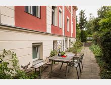 Hinterhofbereich eines Wohnhauses mit roter und cremefarbener Fassade, gepflastertem Weg und Tischgruppe. Auf dem Tisch liegen eine Wassermelone, Paprika und zwei Weinflaschen, umgeben von vereinzelten Pflanzen und Bäumen im Hintergrund.