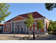 Rotes Kulturzentrum mit Säulenfassade und rotem Dach auf einem gepflasterten Platz, umgeben von Bäumen und Bänken bei sonnigem Wetter.