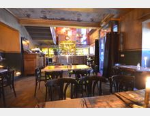 Gemütliches Café mit dunklen Holztischen und Stühlen, einer zentralen Bar im Hintergrund, die von Regalen mit Flaschen und einem beleuchteten Tresen umgeben ist. Die Decke ist mit kunstvollen Malereien verziert, und Kerzen auf den Tischen schaffen eine warme Atmosphäre.