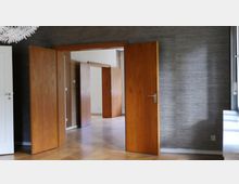 Ein Innenraum mit mehreren geöffneten Holztüren, die in weitere Räume führen. Der Boden besteht aus hellem Parkett, und die Wände sind mit dunkelgrauen Tapeten bedeckt; rechts ist ein Teil eines Fensters sichtbar.