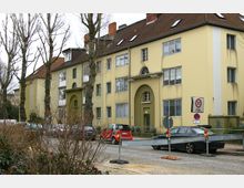 Mehrstöckiges Wohngebäude mit beige-gelber Fassade und braunen Ziegeldächern, das gepflegte, symmetrische Architekturelemente wie gewölbte Eingangsbereiche aufweist. Im Vordergrund stehen parkende Autos entlang einer Pflasterstraße, gesäumt von kahlen Bäumen, sowie Verkehrsschilder, die eine Tempo-30-Zone markieren.