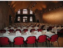 Ein großer, gewölbter Saal mit Backsteinwänden, eingerichtet für ein Bankett mit langen Tafeln, roten Stühlen und gefalteten weißen Servietten. Im Hintergrund befinden sich hohe Fenster mit Rundbögen, die sanftes Licht hereinlassen.