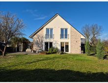 Zweistöckiges Einfamilienhaus mit Satteldach und heller Fassade, teilweise mit Backstein verkleidet. Im Vordergrund ein gepflegter Garten mit Rasen, Bäumen und einer Terrasse auf der linken Seite.