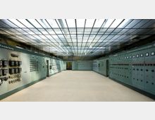 Ein technischer Kontrollraum mit grünen Schalttafeln an beiden Seitenwänden, ausgestattet mit zahlreichen Schaltern, Anzeigen und Messgeräten. Die Decke besteht aus einer großen, gitterartigen Glasstruktur, die den Raum gleichmäßig beleuchtet.