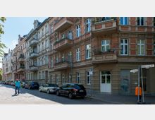 Straßenszene mit Altbau-Wohnhäusern, die durch rote Ziegel und helle Stuckelemente verziert sind. Am Straßenrand stehen geparkte Autos, während ein Fußgänger die gepflasterte Straße entlanggeht; Straßenschilder und ein orangener Mülleimer sind sichtbar.