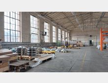 Große Industriehalle mit hohen Decken und sichtbarer Holzkonstruktion, großen Fenstern mit Sprossen und hellem Mauerwerk. Der Boden ist mit Paletten, Maschinen und verpackten Gütern belegt, während Rohrleitungen und Beleuchtung an den Wänden und der Decke sichtbar sind.
