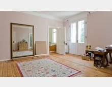 Ein heller Raum mit holzfarbenem Dielenboden, einer großen Wandspiegel mit goldfarbenem Rahmen, einer offenen Doppeltür und einer Glastür, die nach außen führt. Ein farbig gemusterter Teppich liegt auf dem Boden, und im Raum befinden sich Möbel wie ein Schreibtisch, eine Kommode und ein geflochtener Stuhl.