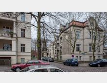 Straßenszene in einer Wohngegend mit gepflasterter Straße, umgeben von mehrstöckigen Altbauwohnungen mit hellen Fassaden und großen Fenstern. Bäume ohne Laub stehen entlang der Straße, auf der mehrere parkende Autos zu sehen sind.