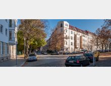 Straßenszene in einem Wohngebiet mit mehrstöckigen Altbauhäusern in heller Fassade und roten Dächern; gepflasterte Straße mit geparkten Autos und herbstlichen Bäumen an den Seiten. Ein Straßenschild mit der Aufschrift „Gürtelstraße“ sowie ein gelber Postbriefkasten sind sichtbar.