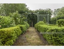 Ein gepflegter Gartenweg führt durch dichte, grüne Hecken und blühende Rosen zu einem dekorativen, grünen Metallgitterbogen, hinter dem eine Statue steht. Im Hintergrund ist dichter Bewuchs mit Bäumen und Sträuchern erkennbar.