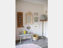 Ein helles Zimmer mit einer cremefarbenen Wand, dekoriert mit einem goldenen Barockspiegel und mehreren kleinen gerahmten Bildern. Ein schmaler Tisch mit Vasen und Dekorationen steht links, daneben ein Korb aus geflochtener Naturfaser und eine antike Stehlampe mit Lampenschirm. Der Boden besteht aus hellgrauem Holz und ein runder Teppich mit rosa Muster liegt davor.