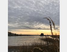 Ein ruhiger See bei Sonnenuntergang mit Schilfgras im Vordergrund. Am Horizont sind ein hölzernes Gebäude auf einem Steg und eine leicht bewölkte Abendhimmel zu sehen.