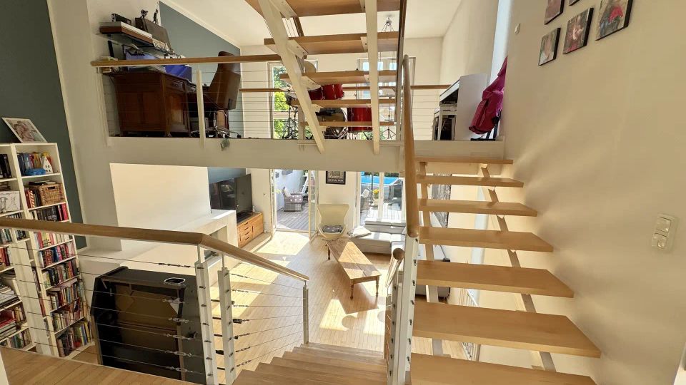 Blick in einen modernen, offenen Wohnraum mit Galerieebene und freitragender Holztreppe mit Metallgeländer. Unten sind ein Wohnzimmer mit Sitzmöbeln, Bücherregal und großen Glas-/Terrassentüren zu sehen, oben eine offene Arbeits- oder Musikfläche mit Möbeln und Instrumenten.