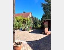 Pflasterstein-Hof mit einem Gebäude aus Stein und einem roten Ziegeldach, teils von Pflanzen bewachsen. Im Vordergrund stehen Blumentöpfe mit Rosen, im Hintergrund sind Bäume und ein Holzzaun zu sehen.