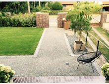 Gepflegter Vorgarten mit gepflastertem Weg, der zu einem Gartentor aus Metall führt, flankiert von niedrigen Backsteinmauern. Rechts neben dem Weg befinden sich Pflanzkübel mit kleinen Bäumen und ein moderner schwarzer Stuhl; links ist eine Rasenfläche.