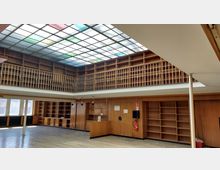 Großer Raum mit Holzregalen an den Wänden und einer umlaufenden hölzernen Galerie im oberen Bereich. Die Decke ist mit einem großen farbigen Glasdach versehen, das Tageslicht hereinlässt, und der Raum verfügt über einen Holzfußboden sowie eine großzügige Fensterfront.