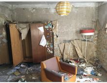 Ein verlassenes Zimmer mit beschädigten Wänden, abblätterndem Tapetenstücken und Unordnung auf dem Boden. Zu sehen sind ein offener Holzschrank, ein orangefarbener Sessel, ein hängender Lampenschirm sowie weitere Gegenstände, darunter ein alter Trockenhaubenständer.